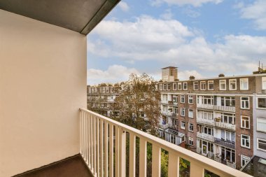 Arka planda bazı binalar ve üzerinde mavi gökyüzü olan bir balkon, bir apartman binasının balkonundan görüldüğü gibi.