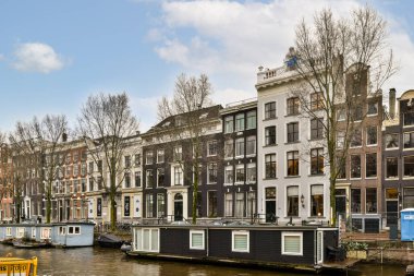 Amsterdam, Hollanda - 10 Nisan 2021: Kanalın diğer tarafındaki bazı tekneler ve üzerinde mavi gökyüzü olan binalar, karşı kıyıdan görüldüğü gibi.