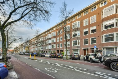 Bir apartman ve iki katlı tuğla bina önünde yolun kenarına park etmiş arabalarla dolu bir şehir caddesi.