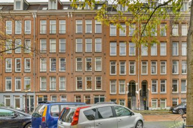 İki araba büyük bir tuğla binanın önüne park etmiş, her iki tarafında da birçok pencere var ve bir arabanın kapısı açık.