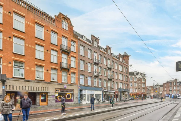 stock image Amsterdam, Netherlands - 10 April, 2021: some people walking down the street in front of an old red brick building on a sunny day with blue sky and white clouds