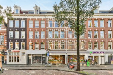 Amsterdam, Hollanda - 10 Nisan 2021: Birçok penceresi olan tuğla bir binanın önündeki ağaç ve bazı binaların yanındaki kaldırımda yürüyen insanlar