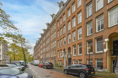 Uzun tuğlalı binalar ve bir sürü penceresi olan kentsel bir alanda caddenin kenarına park edilmiş bazı arabalar var.