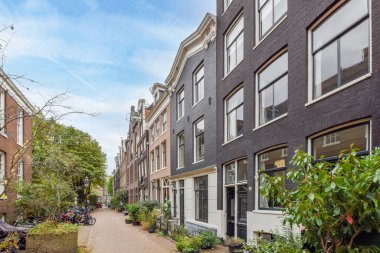 Amsterdam, Hollanda - 10 Nisan 2021: Kenarında park etmiş araçlar olan bir şehir caddesi ve bazı apartmanların önünde kaldırımdan yürüyen insanlar