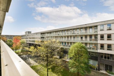 Ön planda binalar ve ağaçlar olan bir dış alan. Balkondan sokağa bakıyorlar.