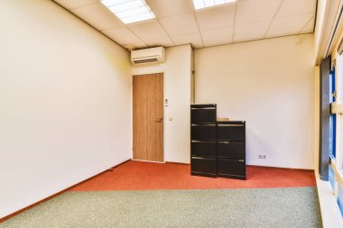 Kırmızı halısı ve beyaz duvarları olan boş bir ofis köşesinde küçük siyah bir dosya dolabı var.