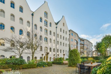 Güneşli bir günde etrafı ağaçlar ve çalılarla çevrili bir apartmanın ortasında masa ve sandalyelerle dolu bir avlu.