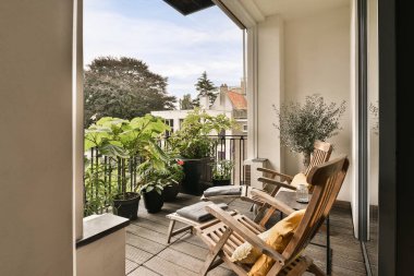 Güvertede ahşap sandalyeler ve saksılarla dolu bir balkon, açık bir kapıdan sokağa bakıyor.