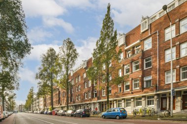 Amsterdam, Hollanda - 10 Nisan 2021: Tuğla binaların önünde yol boyunca ağaçların ve arabaların park ettiği bir şehir caddesi