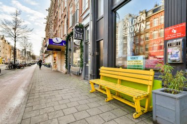 Amsterdam, Hollanda - 10 Nisan 2021: Arka planda binalar ve kaldırımda yürüyen insanlar olan bir caddenin kenarında sarı bir bank