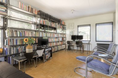 Kitaplarla dolu kitaplığın önündeki raflarda ve sandalyelerde bir sürü kitap olan bir oturma odası.