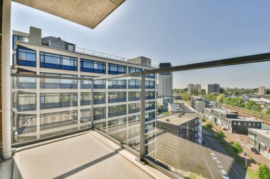 Arka planda bazı binalar olan bir balkon ve ofis penceresinden çekilen şehir manzarasının üzerinde mavi gökyüzü.