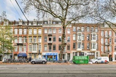 Amsterdam, Hollanda - 10 Nisan 2021: Kenarında arabalar ve arka zemininde yapraklarla kaplı binalar olan bir şehir caddesi