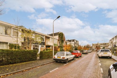 Yan tarafında arabalar ve arka tarafında evler olan bir cadde, bazı binalar görülebilir durumda.
