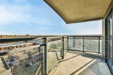 Arka planda bazı binalar ve üzerinde mavi gökyüzü olan bir balkon, bir apartman penceresinden görüldüğü gibi.