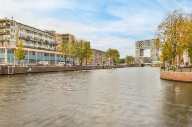 Amsterdam, Hollanda - 10 Nisan 2021: Önünde binalar ve ağaçlar olan bir nehir, arka tarafındaki hareket eden bir tekneden alındı.