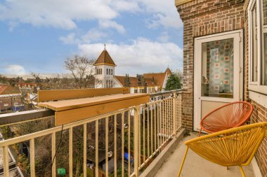 Üzerinde turuncu bir sandalye olan bir balkon ve uzak bir saat kulesi, parlak mavi gökyüzüne karşı.