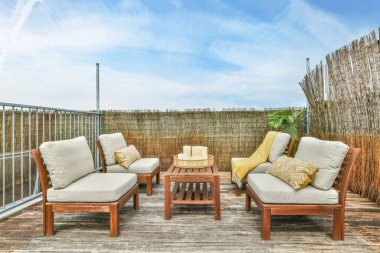 Koltuğun önünde yastıklar ve yastıklarla bir güvertede iki sandalye. Arkalarında mavi gökyüzü beyaz bulutlar.