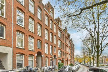 Amsterdam, Hollanda - 10 Nisan 2021: Pencereleri ve balyaları olan tuğla binaların önüne park edilmiş arabalar