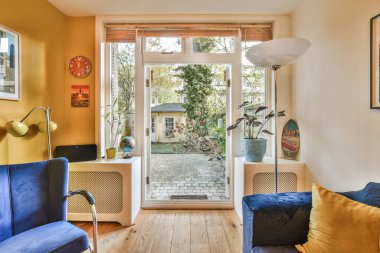 Sarı duvarları ve mavi koltukları olan bir oturma odası arka planda arka bahçeye açılan açık bir kapı var.