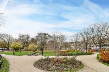Amsterdam, Hollanda - 10 Nisan 2021: Önünde ağaçlar ve banklar olan bir park, açık mavi bir gökyüzü gününde beyaz bulutlarla kaplanmış.