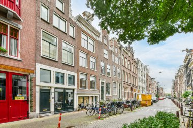 kırmızı pencereli ve yeşil ağaçlı binaların önündeki caddenin kenarına parketmiş birkaç bisiklet.