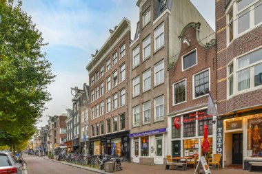 Amsterdam, Hollanda - 10 Nisan 2021: kırmızı tuğlalı bir binanın önünde yol kenarına park edilmiş binalar ve bisikletlerle dolu bir şehir caddesi