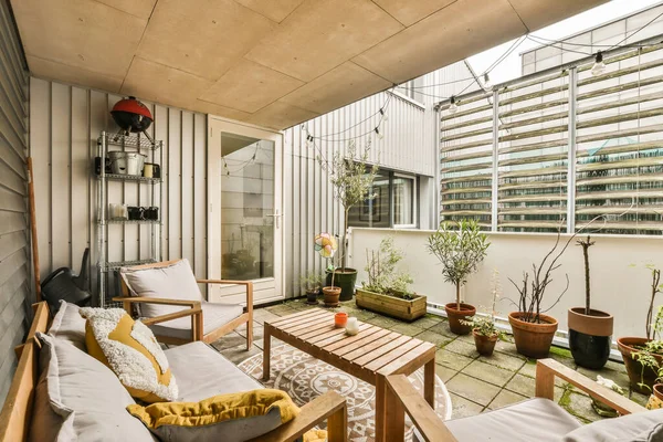 Dış duvarında mobilya ve saksı bitkileri olan bir balkon. Büyük cam bir pencerenin önünde.