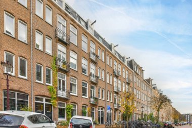 Amsterdam, Hollanda - 10 Nisan 2021: Kenarında arabalar olan bir şehir caddesi ve bazı tuğla binaların önünde yürüyen insanlar
