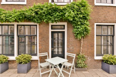 Duvarın arkasında büyüyen bitkilerin olduğu tuğla bir binanın önünde iki sandalye ve bir masa.