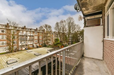 Arka planda bazı binalar olan bir balkon ve üzerinde beyaz bulutlarla kaplanmış mavi bir gökyüzü.