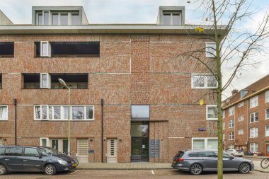 İkinci katta açık penceresi olan tuğla bir binanın önüne park edilmiş iki araba ve yanında da bir araba var.