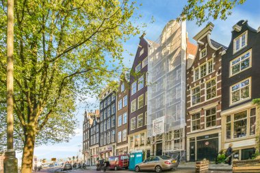 Amsterdam, Hollanda - 10 Nisan 2021: Kenarında arabalar olan bir şehir caddesi ve binaların, ağaçların önünde yürüyen insanlar