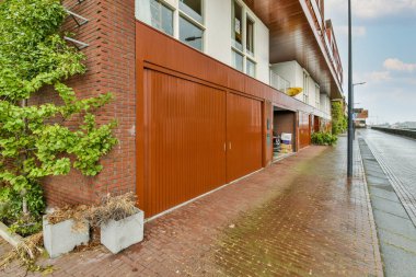 Turuncu garaj kapısının yanında ağaçların ve bitkilerin yetiştiği kırmızı tuğlalı bir bina.