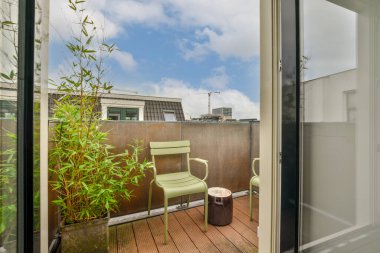Bir balkon, bir sandalye ve yerde saksı bitkisi. Açık cam bir kapının önünde.