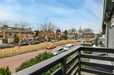 Kaldırımda arabaların park ettiği ve uzaktaki evlerin olduğu bir cadde. Dışarıdaki bir balkondan sokağa bakıyorlar.