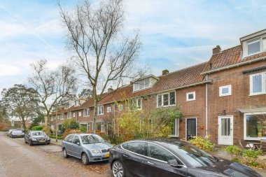 Amsterdam, Hollanda - 10 Nisan 2021: Bazı arabalar eski bir tuğla binanın önündeki caddenin kenarına park edilmiş ağaçlar ve çalılar boyunca büyüyor.