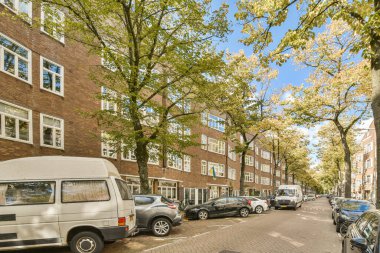 Bazı arabalar sokağın kenarına park edilmiş tuğla bir binanın önünde, etrafında birçok pencere ve ağaç var.