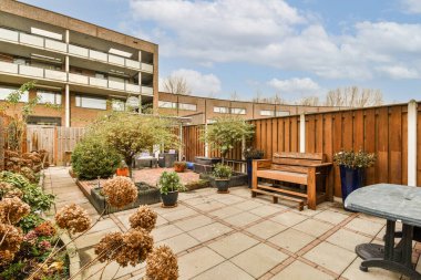 Ağaçlarla çevrili tuğla bir binanın önünde bazı bitki ve çiçeklerin bulunduğu bir alan.