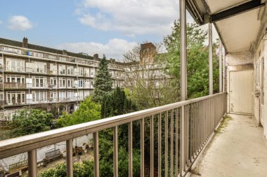Arka planda ağaçlar ve binalar olan bir balkon. Bir apartmanın penceresinden sokağa bakıyor.