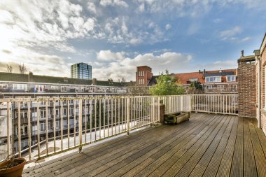 Ağaçlarla çevrili tuğla bir binanın kenarındaki ahşap döşemeli ve beyaz korkuluklu bir alan.