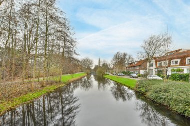 Amsterdam, Hollanda - 10 Nisan 2021: Güneşli bir günde arka planda evleri ve ağaçları olan bir kanal, deniz kıyısından görüldüğü gibi.