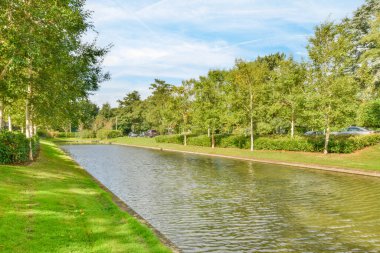 Amsterdam, Hollanda - 10 Nisan 2021: Her iki tarafında ağaçlar ve çimenler olan bir parkın ortasındaki kanal boyunca bazı arabalar park halindedir.