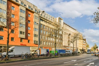 Turuncu tuğlalı bir binanın önünde yol kenarına park etmiş arabaları ve bisikletleri olan bir cadde.