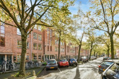 Amsterdam, Hollanda - 10 Nisan 2021: Mavi gökyüzünün altında kırmızı tuğlalı binaların önündeki kaldırımda park etmiş arabaları ve bisikletleri olan bir şehir caddesi