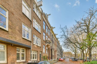 Kaldırımlarda arabaları ve tuğlalı apartmanların önündeki yaprakları olmayan ağaçları olan bir şehir caddesi.