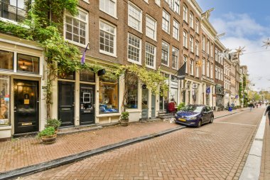 Amsterdam, Hollanda - 10 Nisan 2021: Kenarında arabaları park edilmiş bir şehir caddesi ve tuğlalı binaların önünde yürüyen insanlar