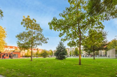 Ağaçlı bir park ve uzaktaki kaldırımda yürüyen insanlar inşa edilmiş kırmızı tuğlalı bir bina.
