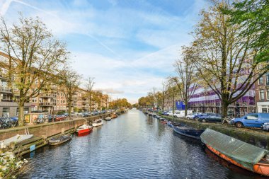 Amsterdam, Hollanda - 10 Nisan 2021: Suda tekneleri ve arka planda evleri olan bir kanal, nehre bakan bir köprüden alındı