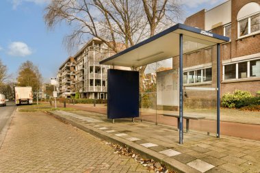 Caddenin kenarında bir otobüs durağı. Arkasında ağaçlar ve binalar olan bir binanın önünde.,